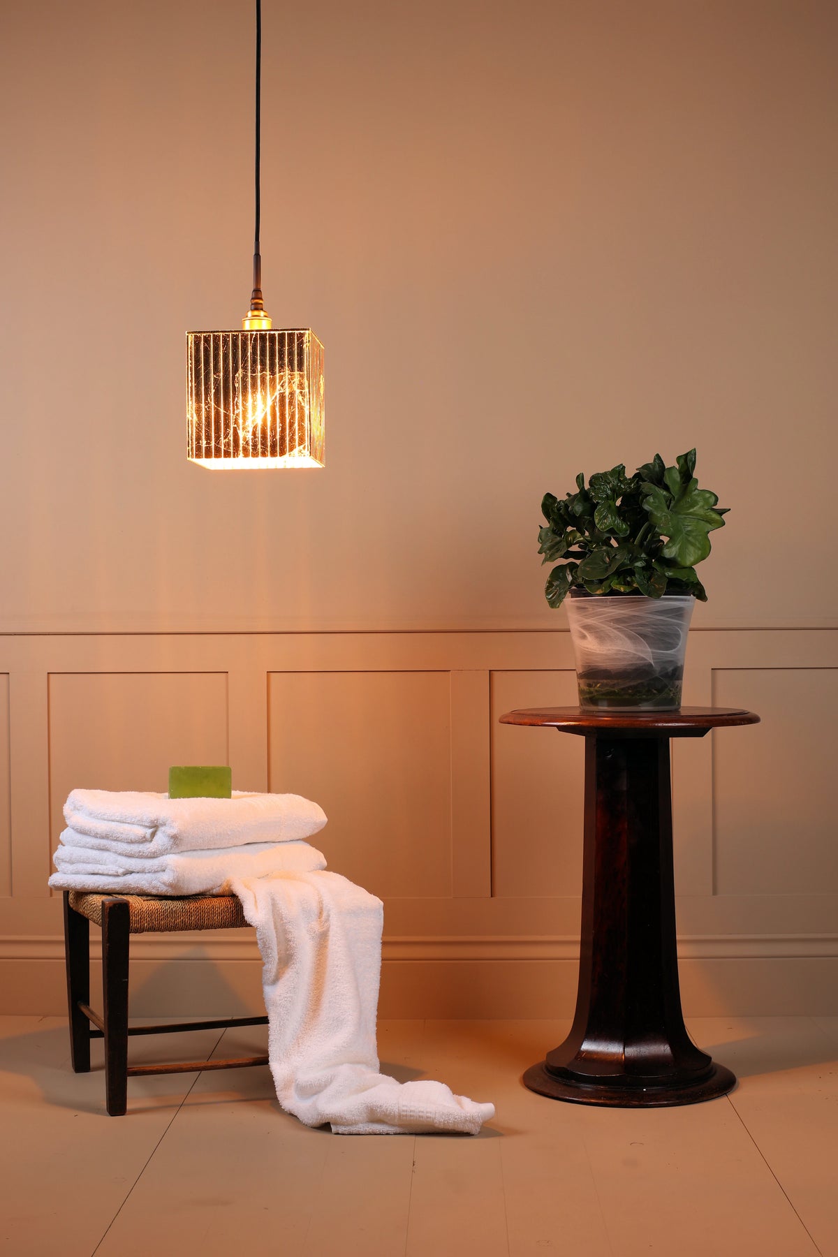 Gold square bathroom pendant light hanging over white towels, wooden table with plant, beige coloured wall