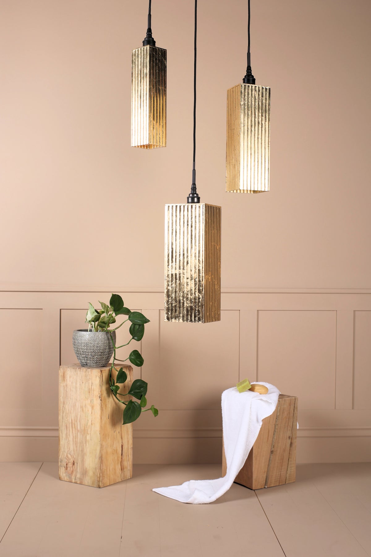 Three modern cylindrical bathroom pendant lights with ribbed metallic gold shades hang against a beige wall with decorative wood blocks and a potted plant.