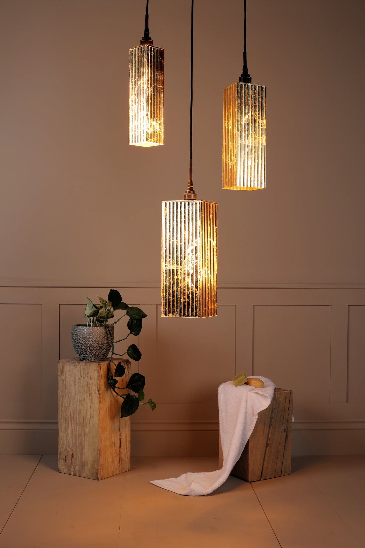 Three cylindrical bathroom pendant lights with textured, ribbed glass shades glowing with a warm golden light, hanging in a styled room setting with wood block pedestals and a draped white cloth.
