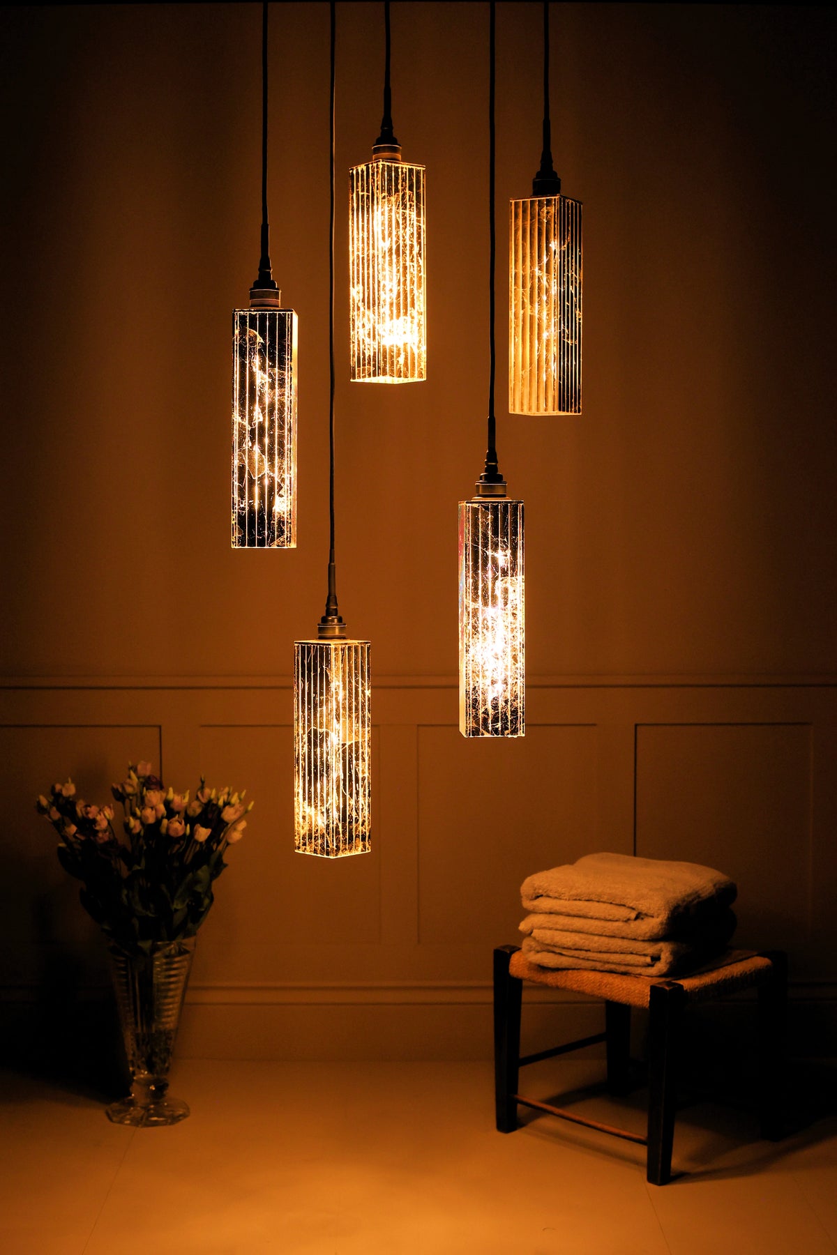 Five modern cylindrical pendant lights with textured, glowing glass shades hanging in a warm, dimly lit room. A small wooden stool with folded towels and a vase of flowers sit below the lights against a dark paneled wall.