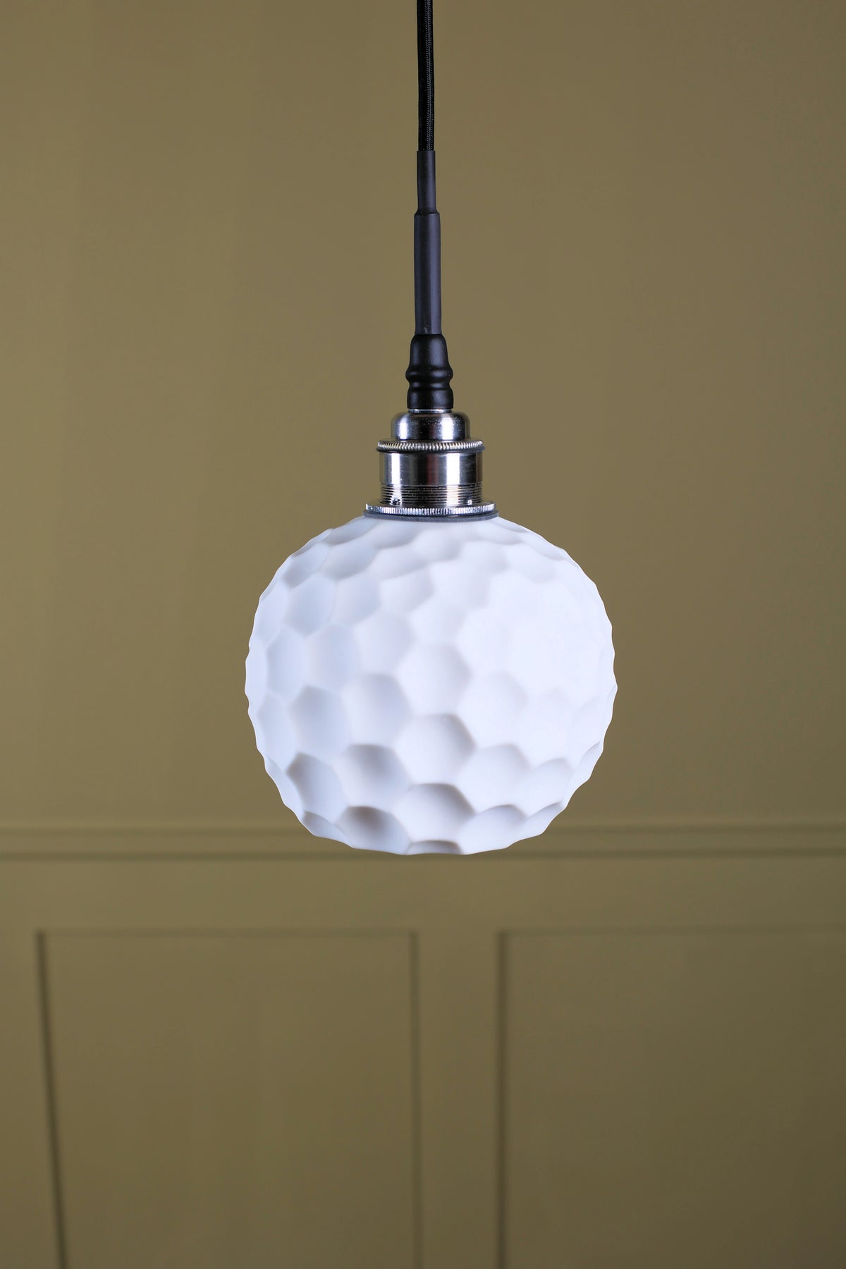 A white textured pendant light shaped like a golf ball, suspended from a black cord with a silver metal collar, set against a warm beige paneled wall.