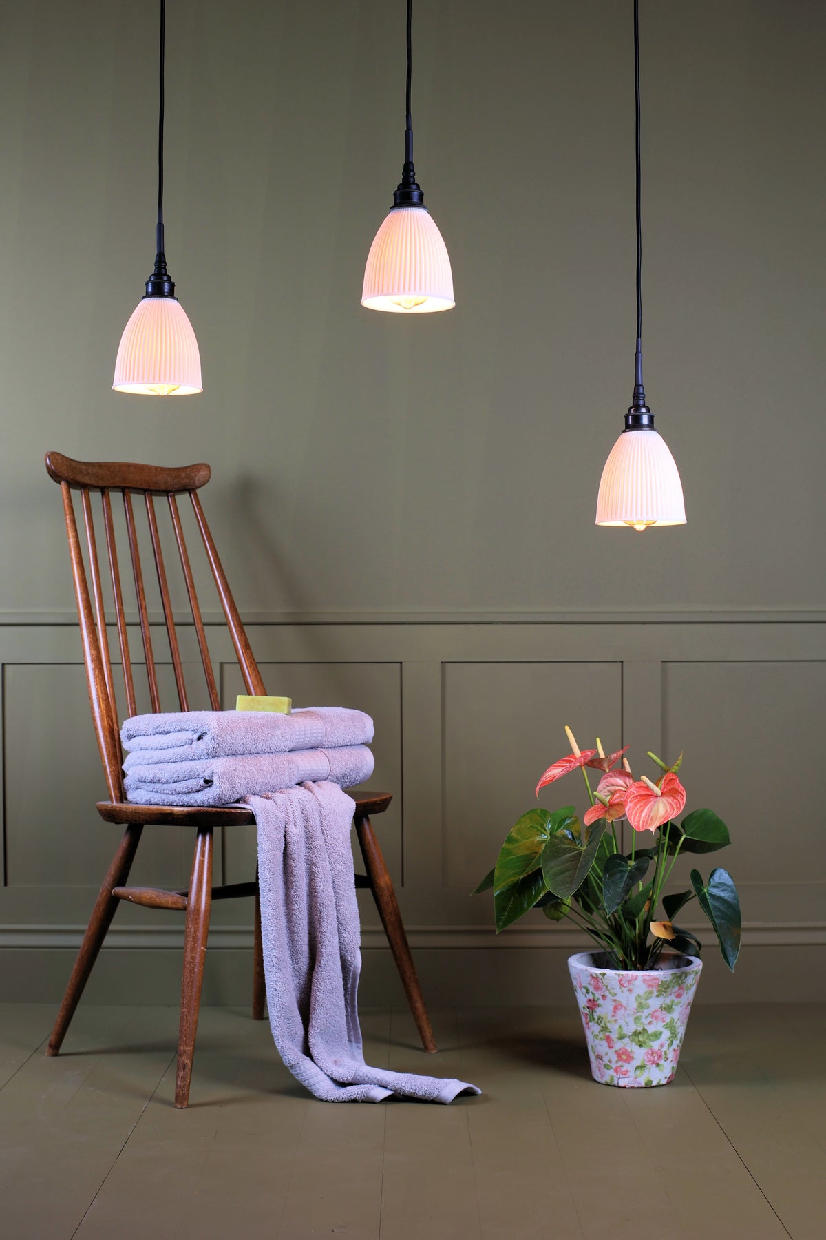 A cluster of three small ceramic bathroom pendant lights, traditional wooden chair, folded bath towels, exotic plant on sage green paneled background