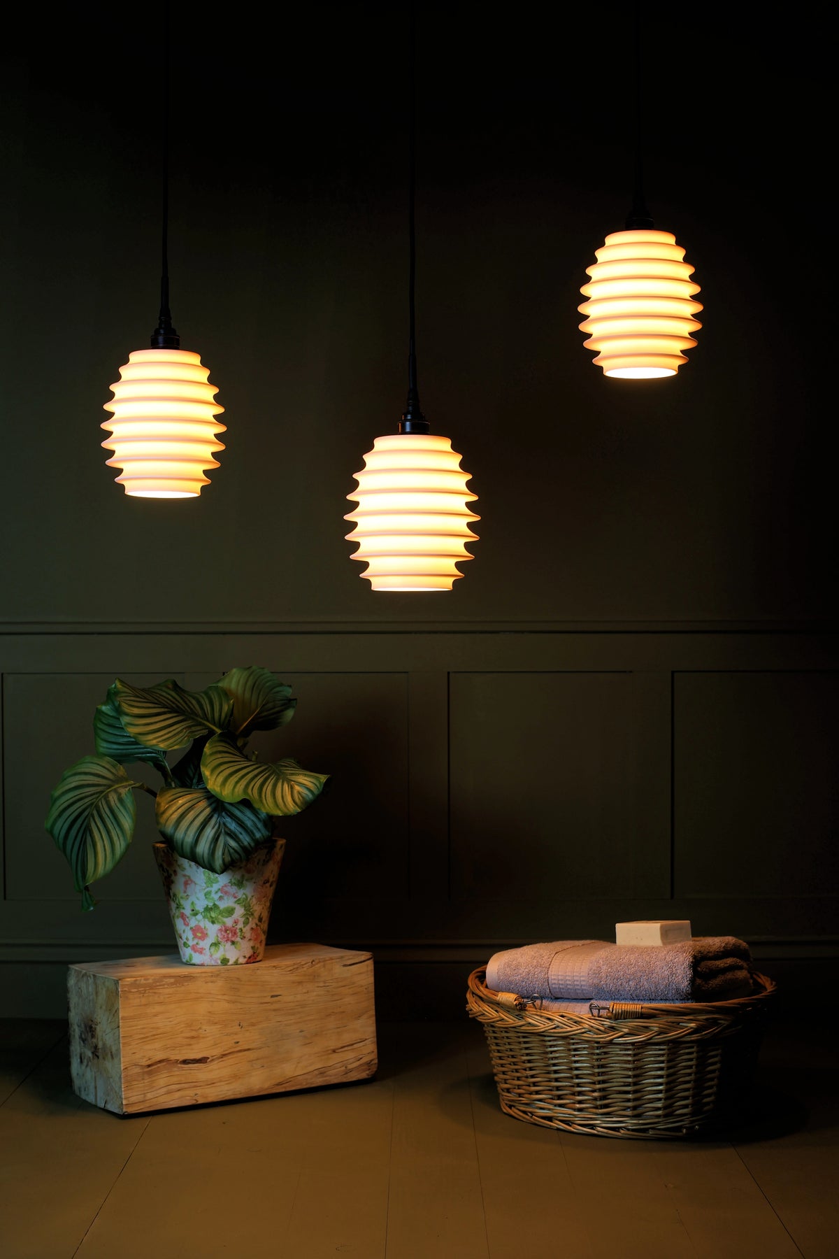 Concertina style porcelain triple cluster bathroom pendant lights with plant and basket of bath towels on sage green background in dimly lit room