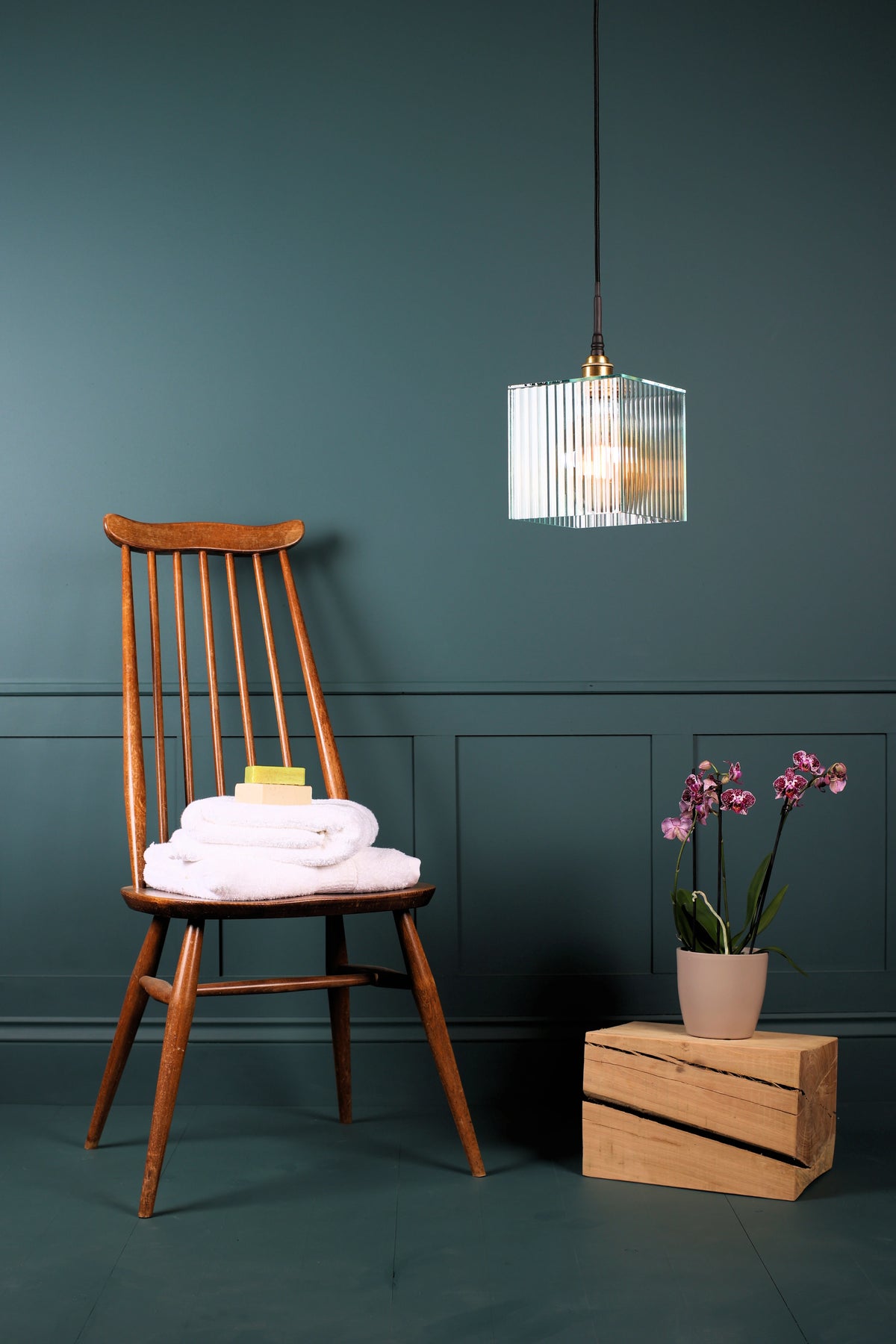 A square ribbed glass bathroom pendant light, antique wooden chair, white bath towels, exotic plant on oak block, teal walls.