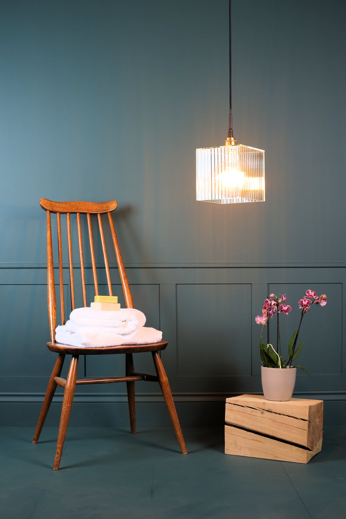 A square ribbed glass bathroom pendant light with warm glowing light bulb, antique wooden chair, white bath towels, exotic plant on oak block, teal wall background