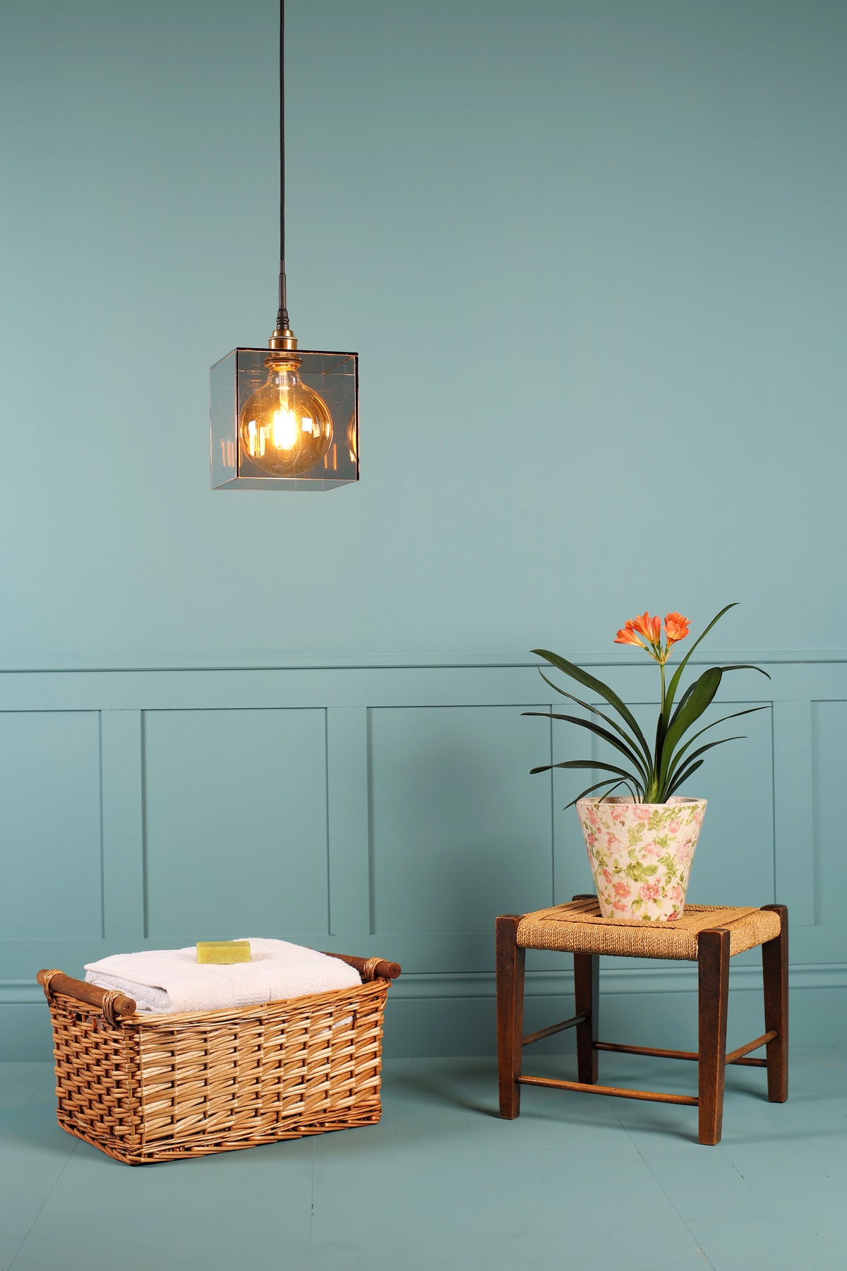 A modern square pendant light with a clear glass shade and warm Edison bulb hanging in a styled room with a blue wall, next to a wicker basket and a small stool with a potted plant.