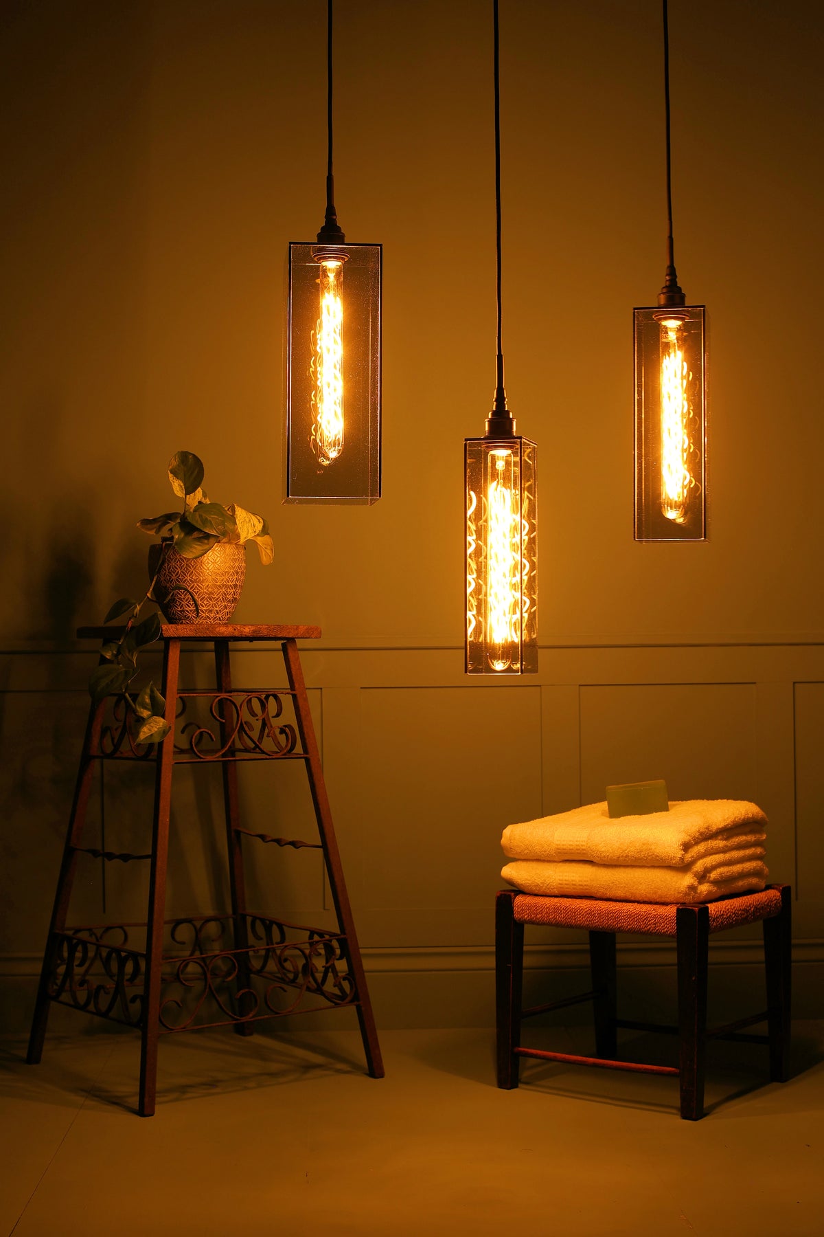 Three rectangular bathroom pendant light fixtures with glowing filament bulbs hanging in a warm, dimly lit room. A small wooden stool with folded towels sits below on the right, and a plant on a metal stand is on the left.