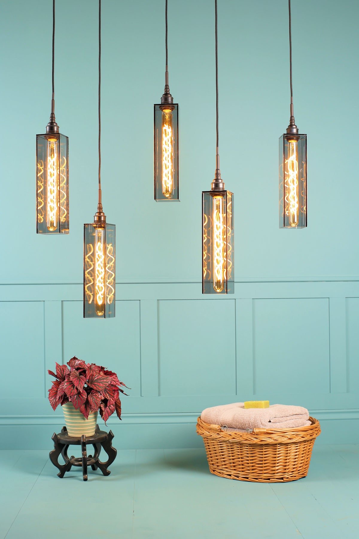 Five industrial-style pendant lights with smoked glass cylinders and glowing Edison bulbs hang against a light blue paneled wall above a floor with a potted plant and a wicker basket.