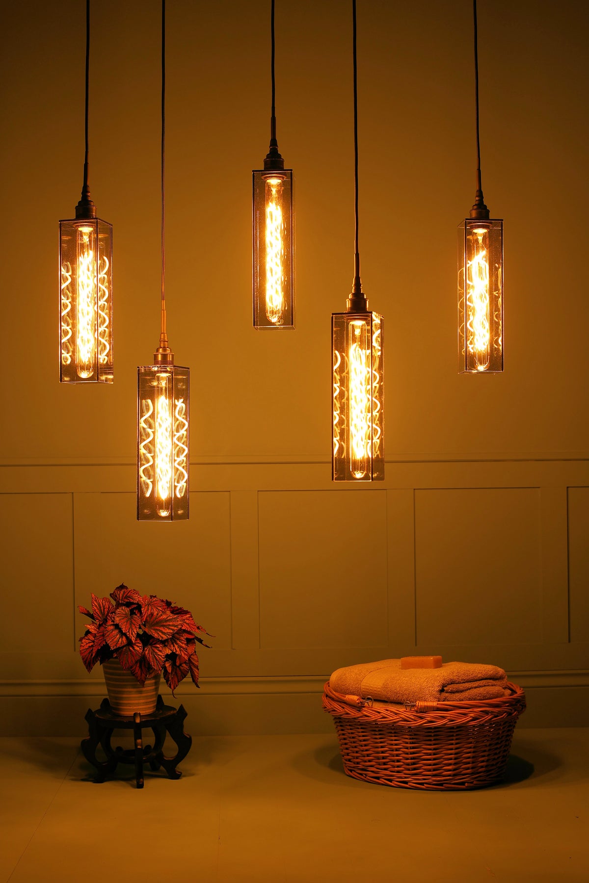Five industrial-style pendant lights with smoked glass cylinders and glowing Edison bulbs hang against a light blue paneled wall above a floor with a potted plant and a wicker basket.