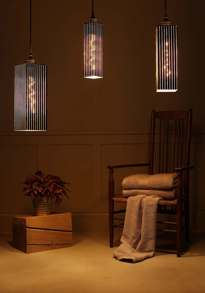 Triple black and gold bathroom pendant light cluster casting shadows onto plant, wooden chair and bath towels.