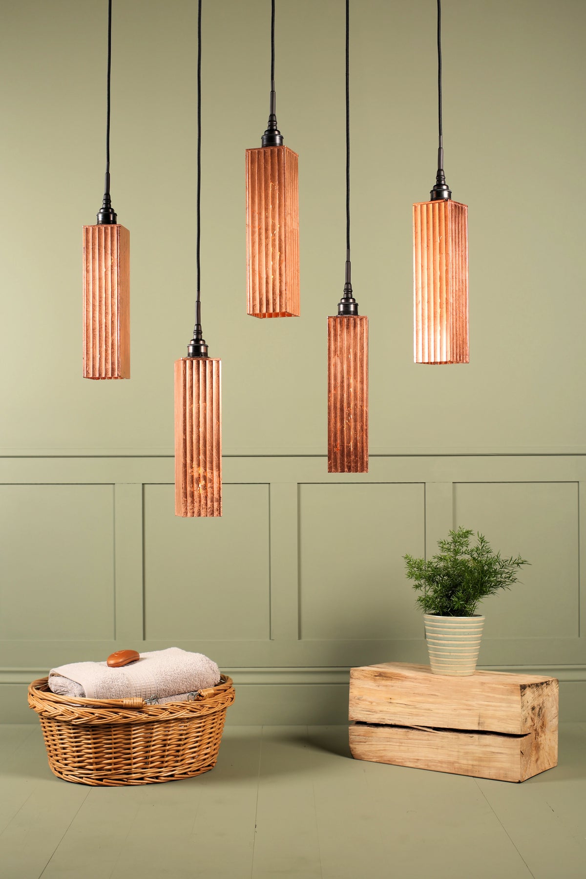Five modern pendant lights with ribbed copper-colored cylindrical shades hanging against a sage green paneled wall. Below, a wicker basket with a folded towel and a small potted plant sit on a wooden block.