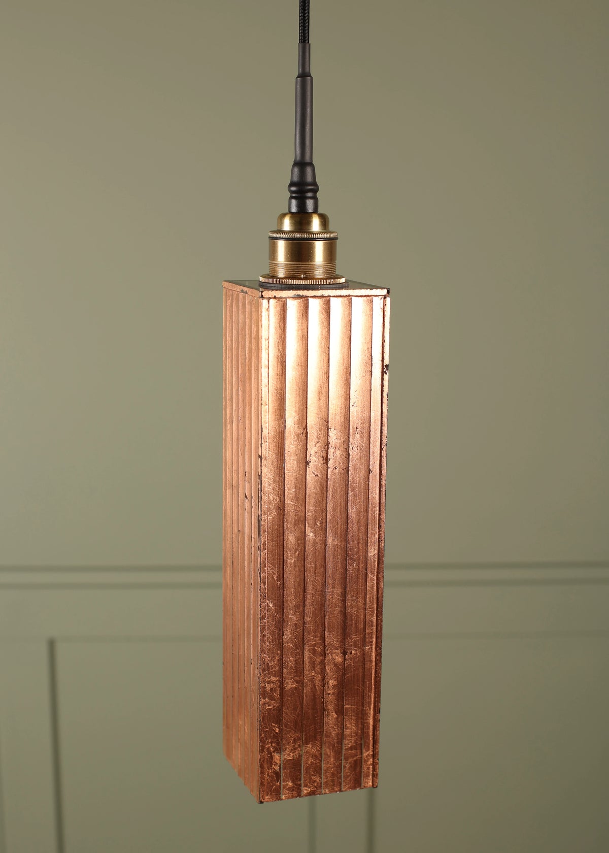 A modern copper bathroom pendant light with a textured, vertically fluted rectangular shade with a brass lamp holder suspended on a black cord on a green paneled background
