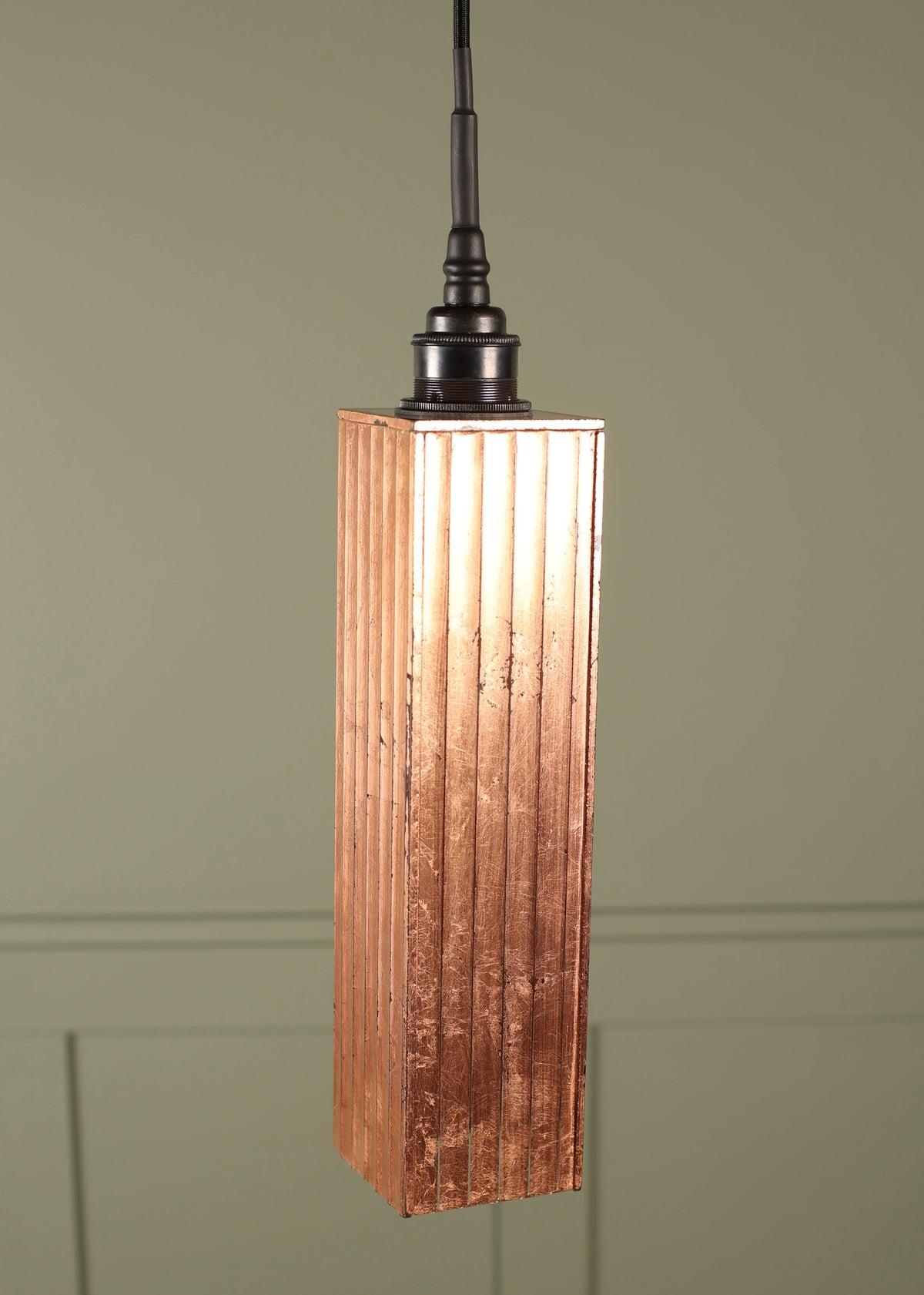 A modern copper bathroom pendant light with a textured, vertically fluted rectangular shade with a dark bronze lamp holder suspended on a black cord on a green paneled background