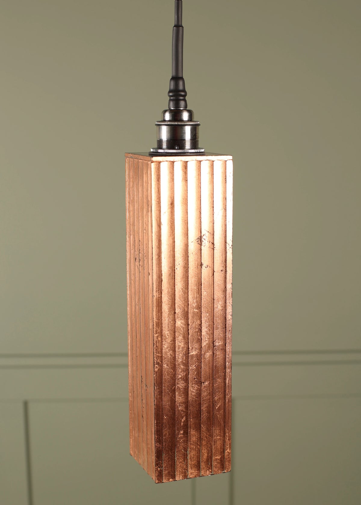 A modern copper bathroom pendant light with a textured, vertically fluted rectangular shade with a chrome lamp holder suspended on a black cord on a green paneled background