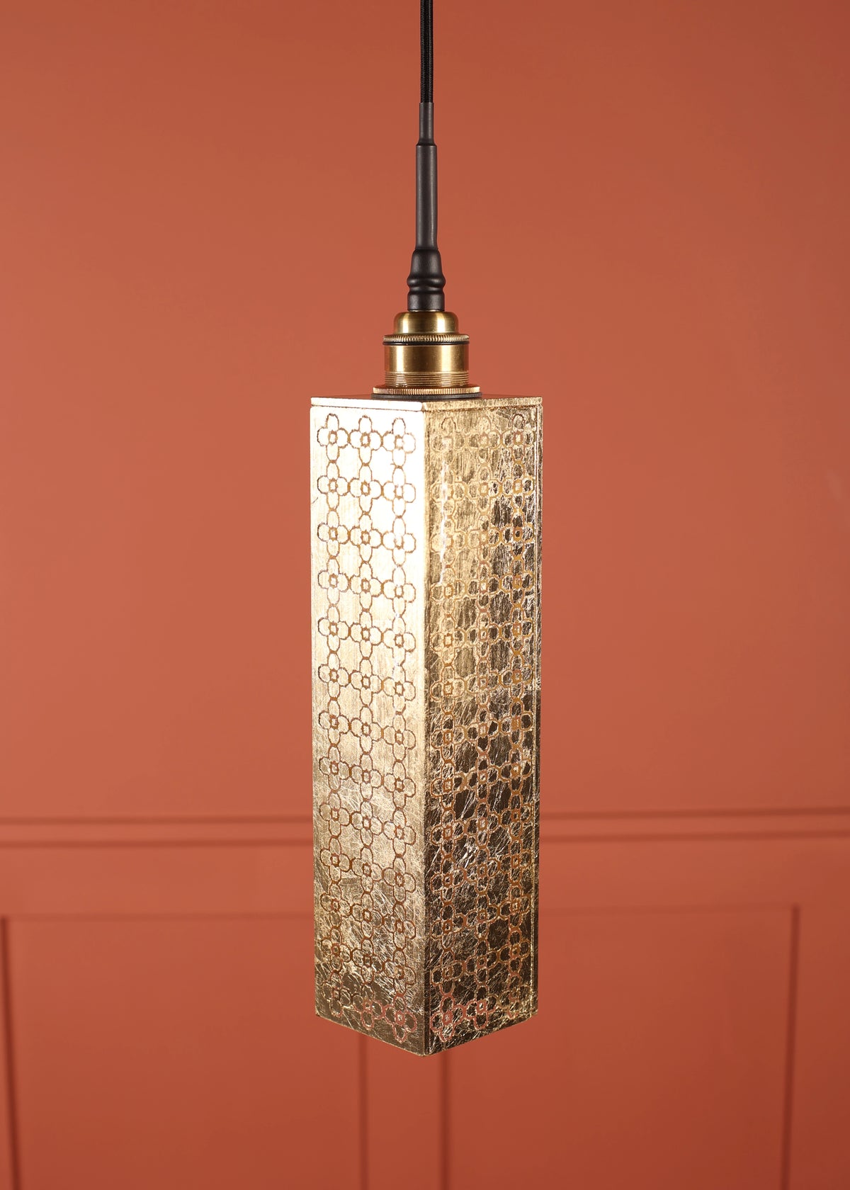 A modern rectangular pendant light with a hammered gold metal shade featuring a geometric trellis pattern, suspended against a terracotta-colored paneled wall.