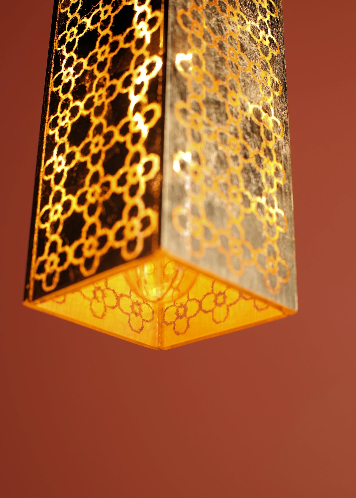 A close-up of a hanging pendant light with a geometric cutout pattern on a textured glass shade, glowing warm yellow against a solid terracotta background.