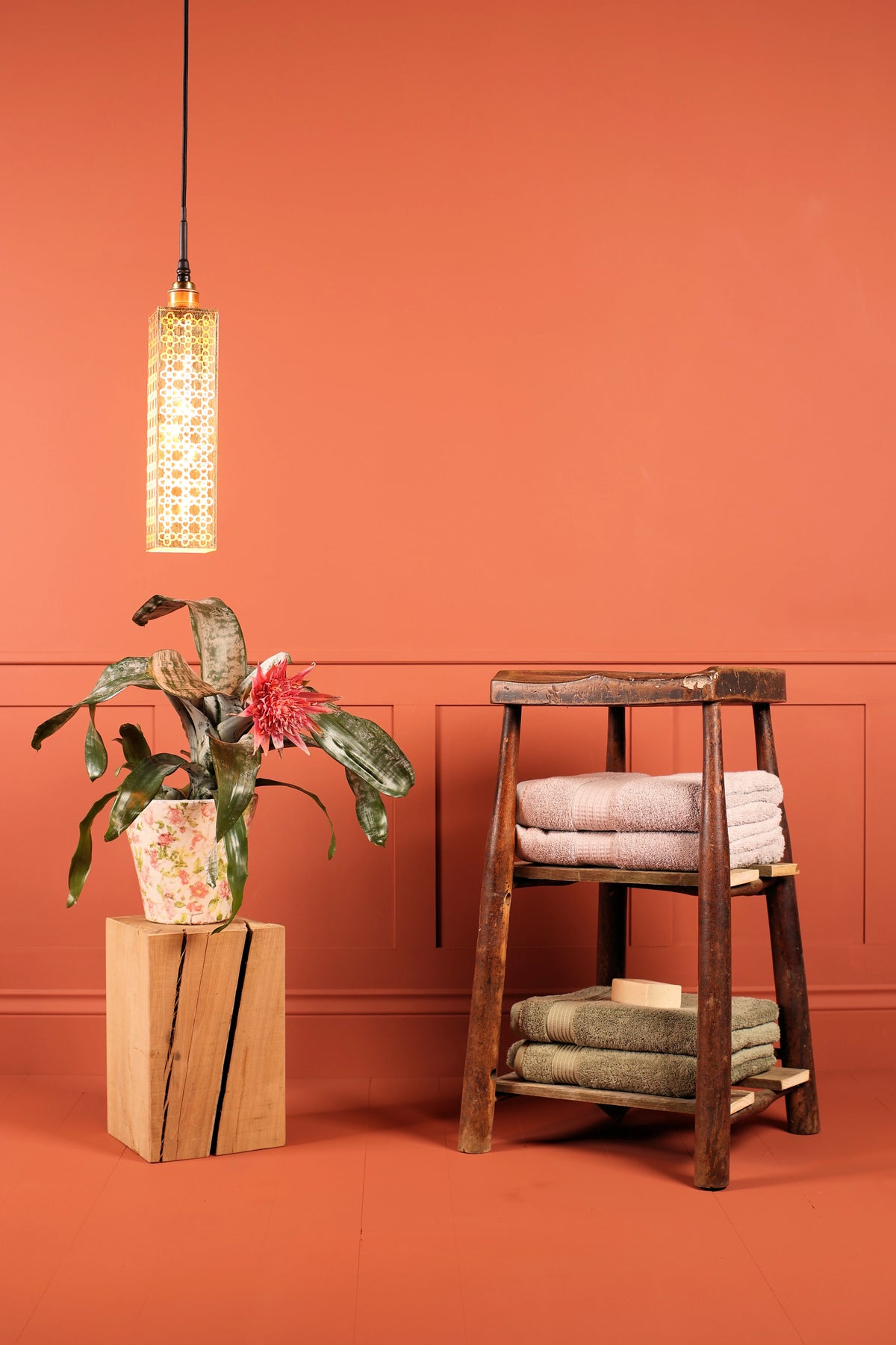 A warm terracotta orange room styled with a rustic wooden three-tier stool holding folded towels, a potted plant on a wood block pedestal, and a hanging gold leaf bathroom pendant light against a paneled wall.