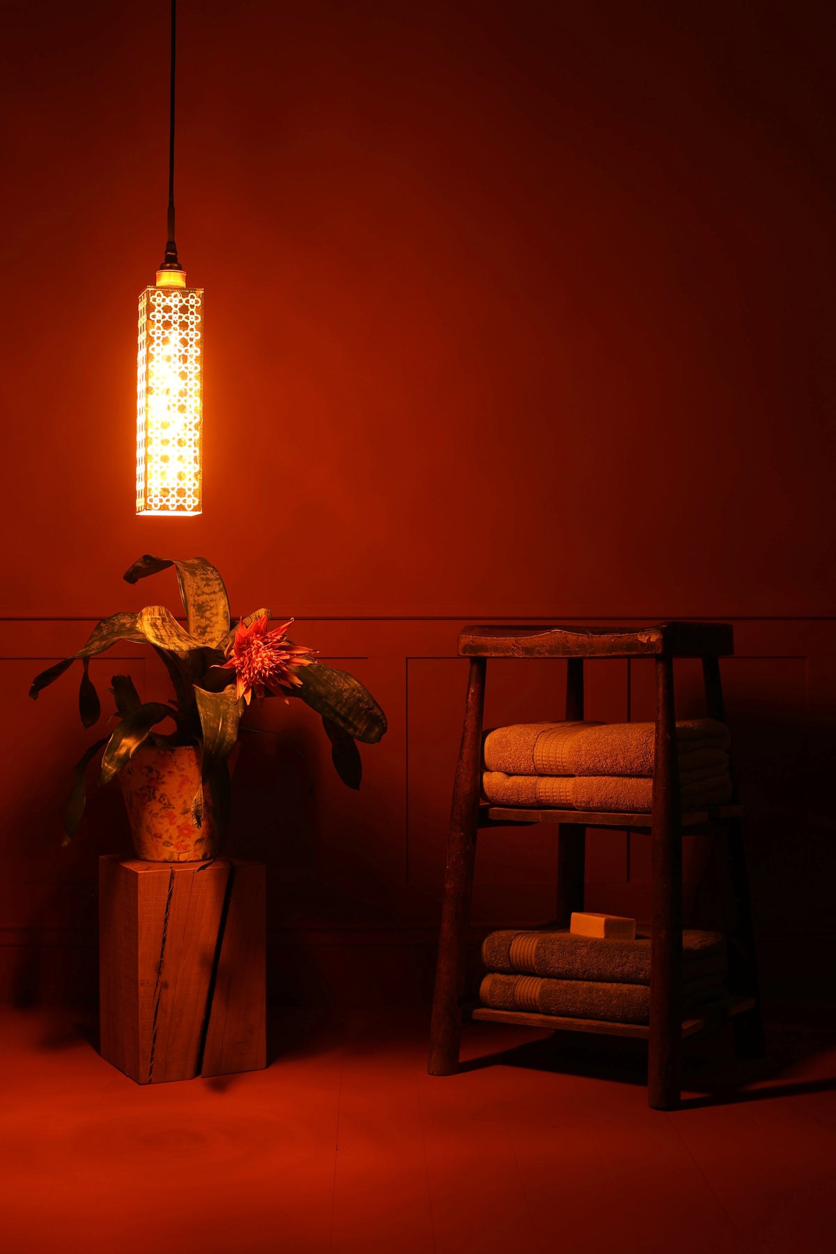 A warm, glowing cylindrical gold leaf bathroom pendant light with a geometric cutout pattern hangs in a dimly lit room with a dark red wall. Below, a wooden stool holds folded towels, and a potted plant sits on a wooden block to the left.