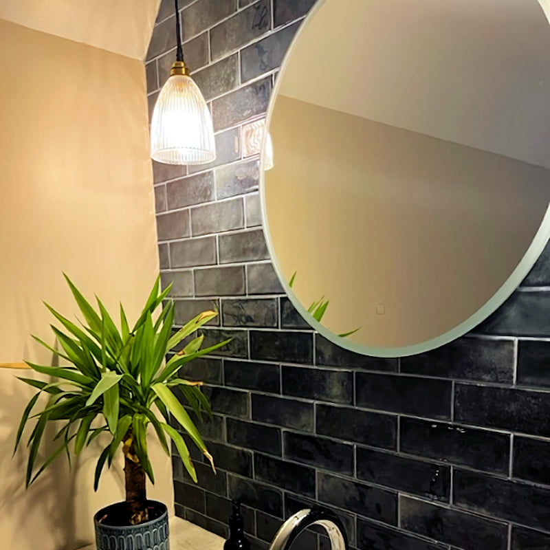 Round mirror on a tiled wall with a plant and glass light fixture in the foreground
