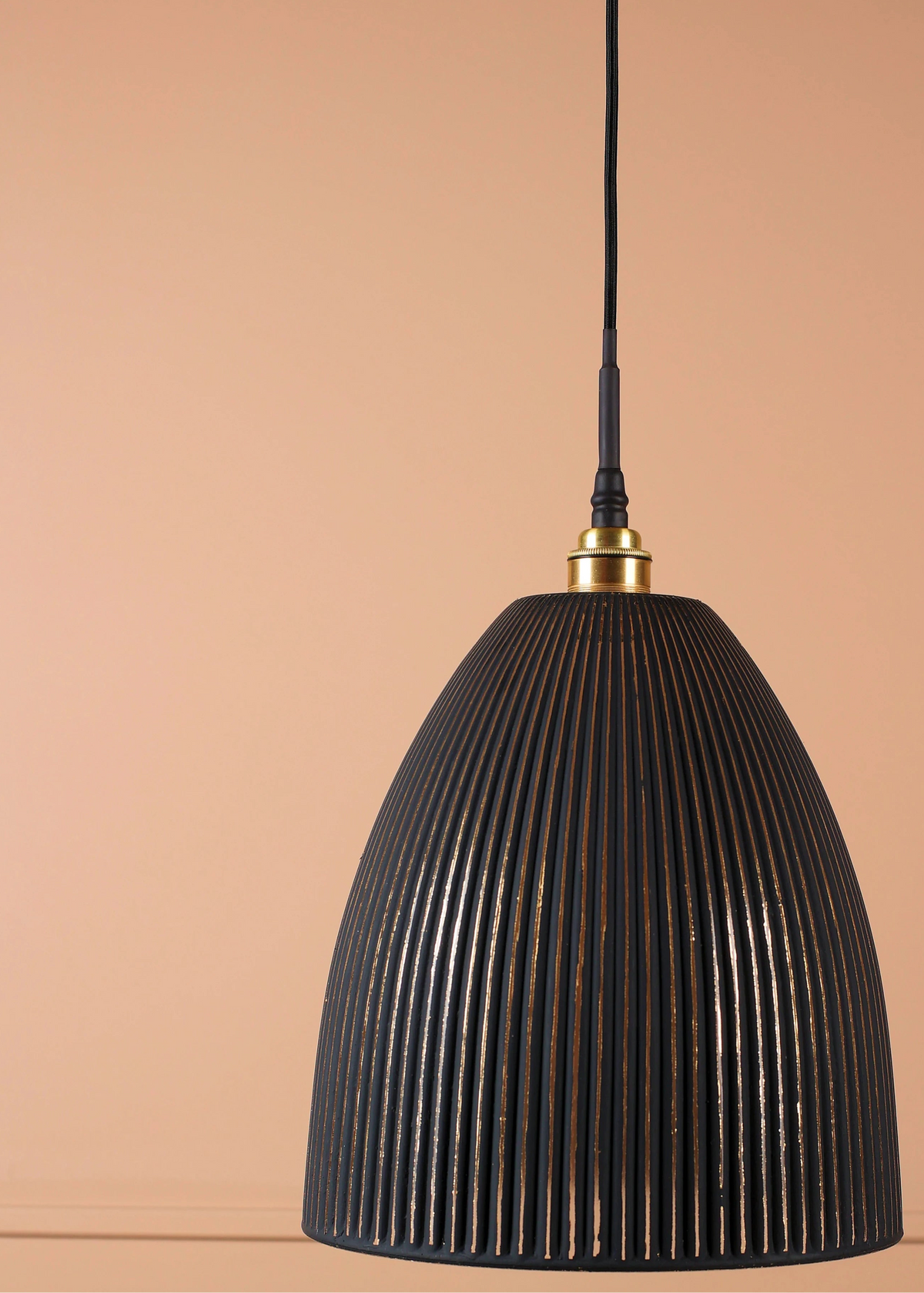A black ribbed conical pendant light hanging from a black cord with a brass fitting, set against a solid peach-colored background.