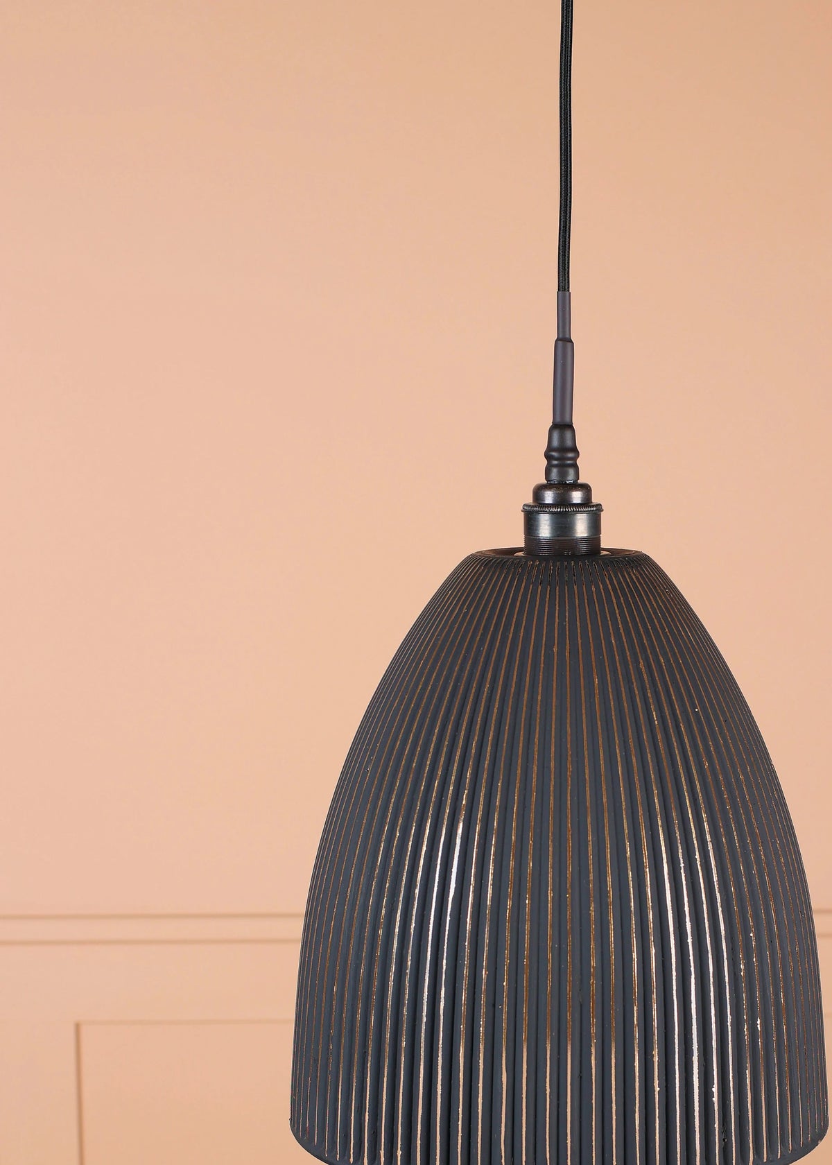 A dark gray ribbed metal pendant light hanging from a black cord against a peach-colored paneled wall.