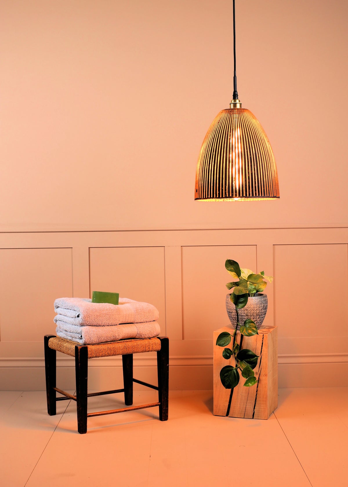 A modern pendant light with a ribbed, fluted metallic dome shade hangs in a warm peach-toned room above a small stool with folded towels and a plant on a wooden block.