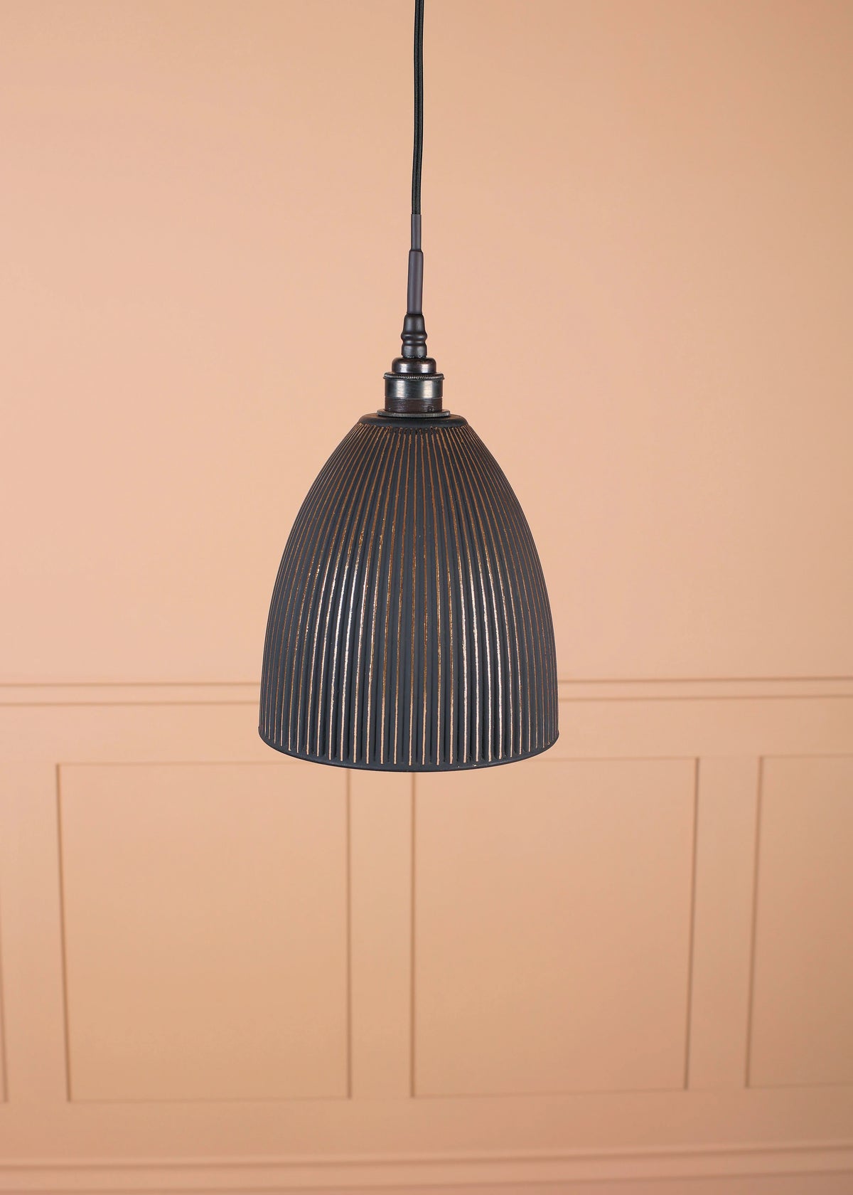 A black ribbed dome pendant light hanging from a black cord against a warm peach-colored wall with decorative panel molding.
