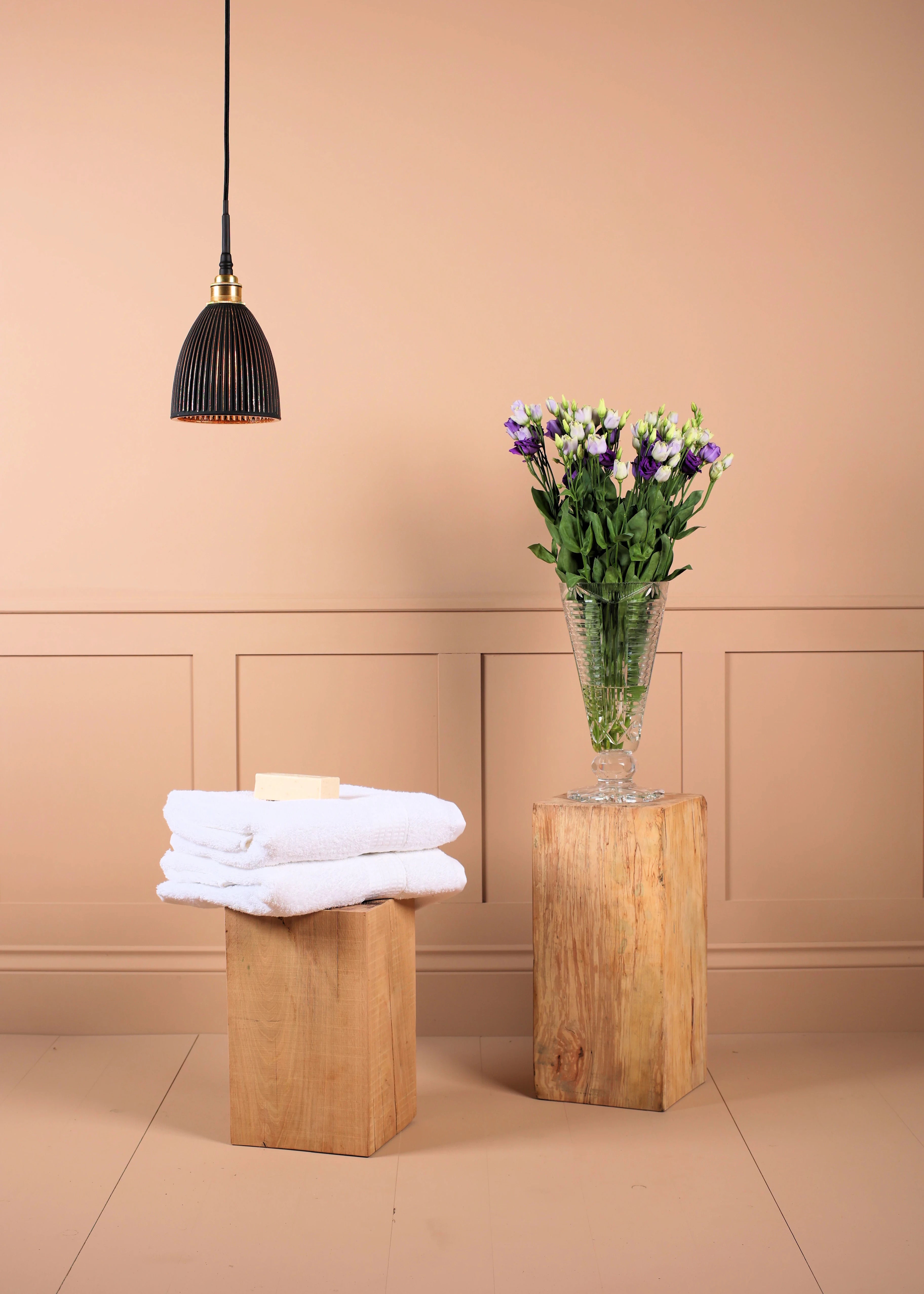 Black & gold bell shaped bathroom pendant light, stack of white towels on a wooden stool with a vase of flowers in the background.