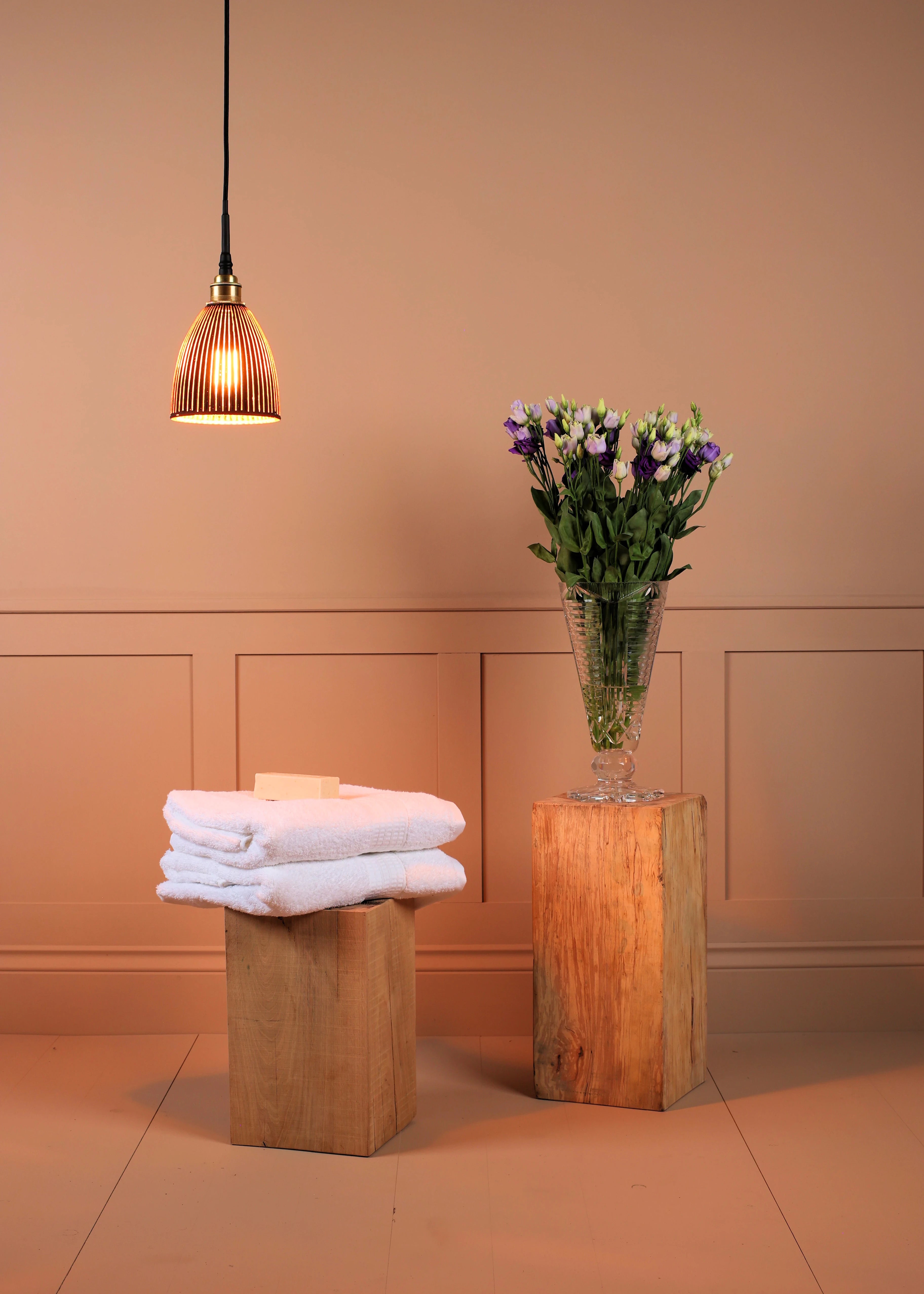 Black & gold bell shaped bathroom pendant light, stack of white towels on a wooden stool with a vase of flowers in the background.