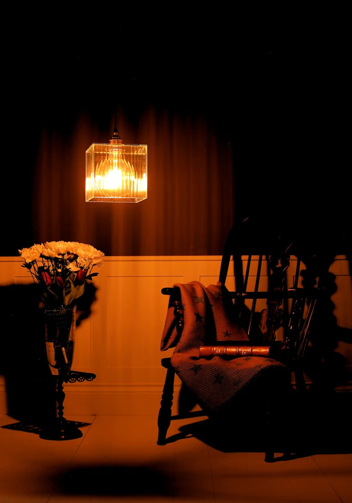 Dark room with a lit lamp, chair, and flowers on a table.