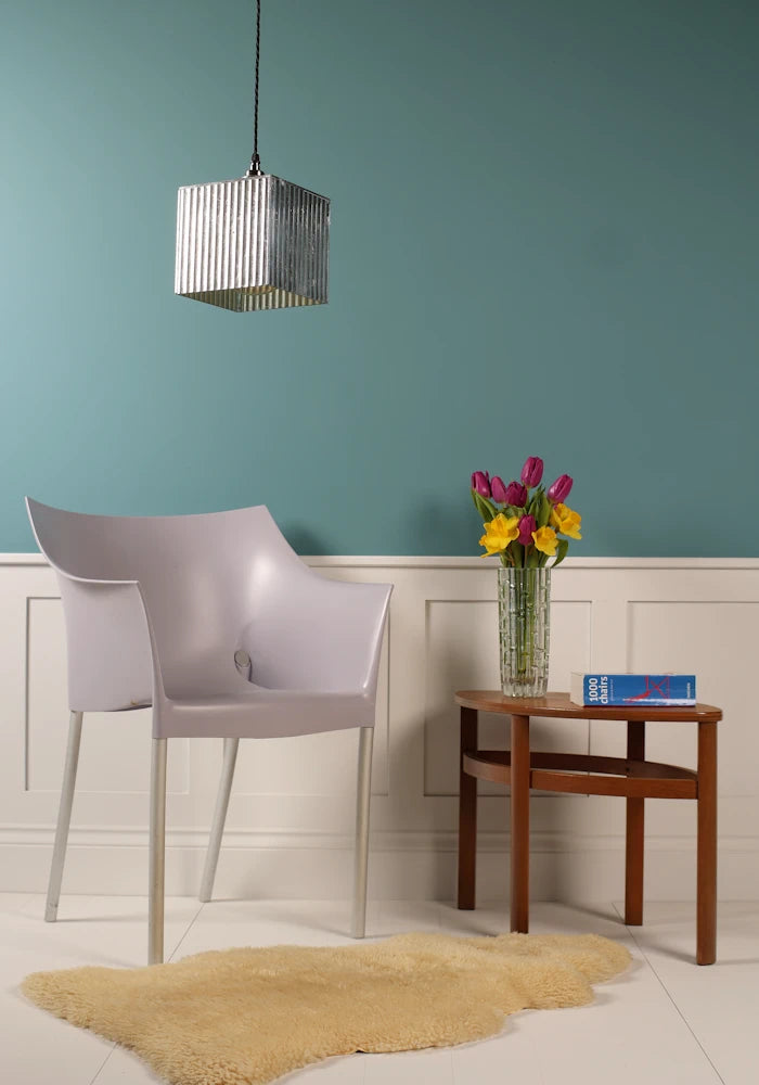 Cubed silver decorative pendant light hanging over modern chair, coffee table with vase of flowers, teal background