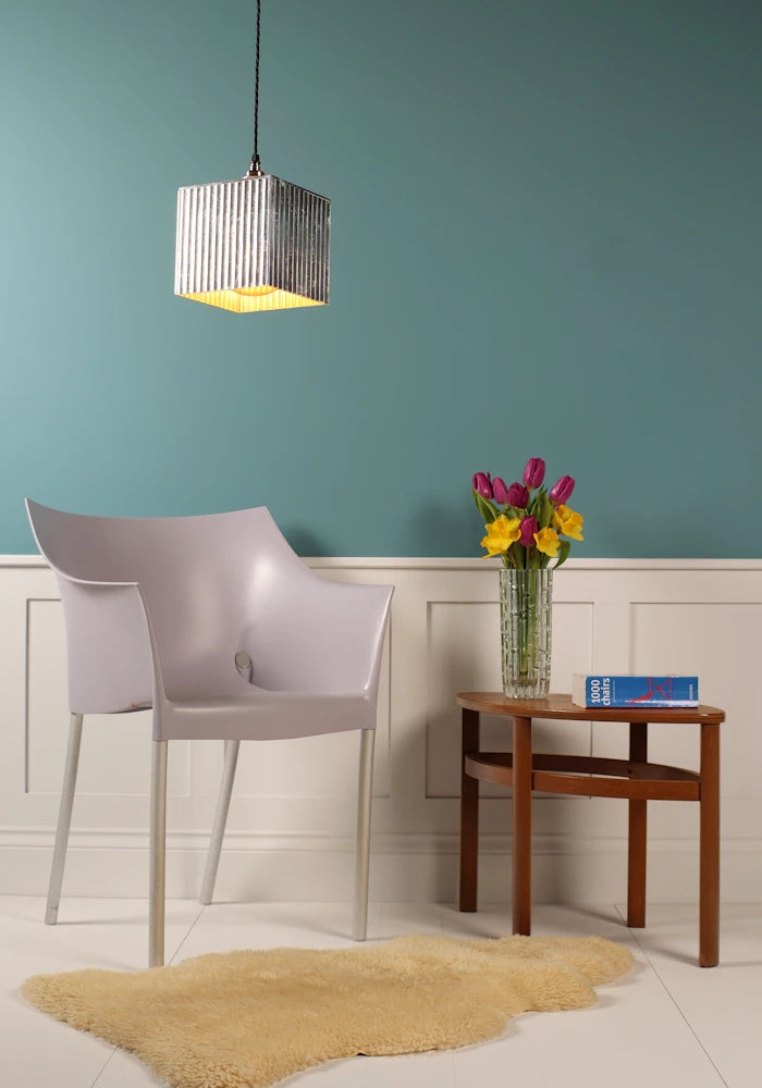 Cubed silver decorative pendant light hanging over modern chair, coffee table with vase of flowers, teal background