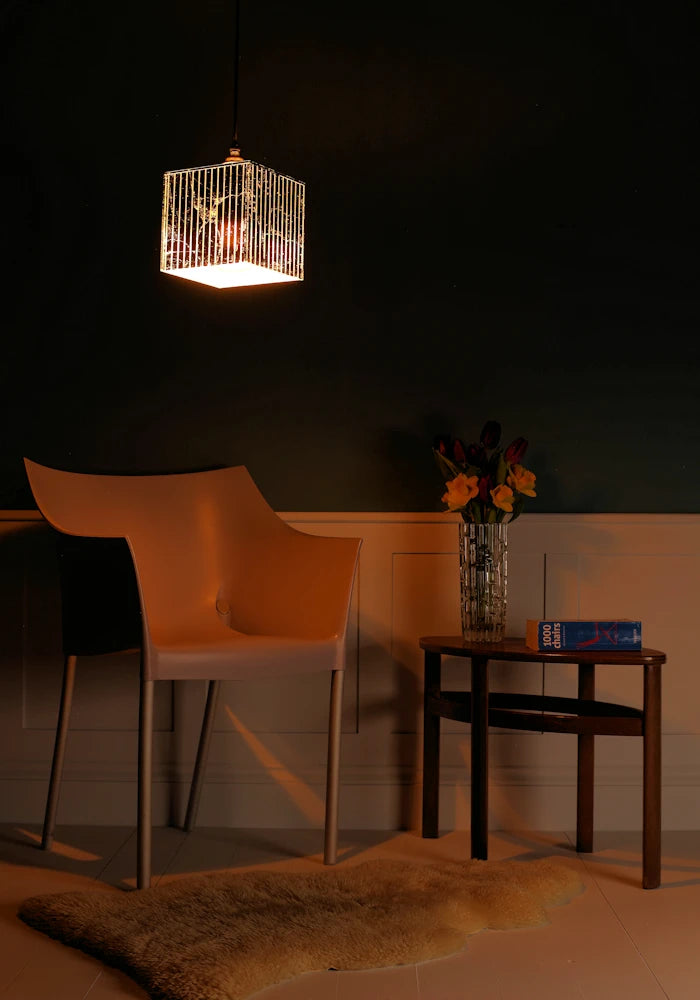 Cubed silver decorative pendant light hanging over modern chair, coffee table with vase of flowers, in dimly lit room