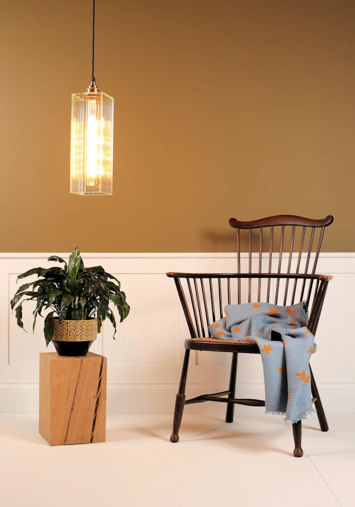 Pendant light fixture, wooden chair with a blanket, and potted plant against a mustard yellow wall.
