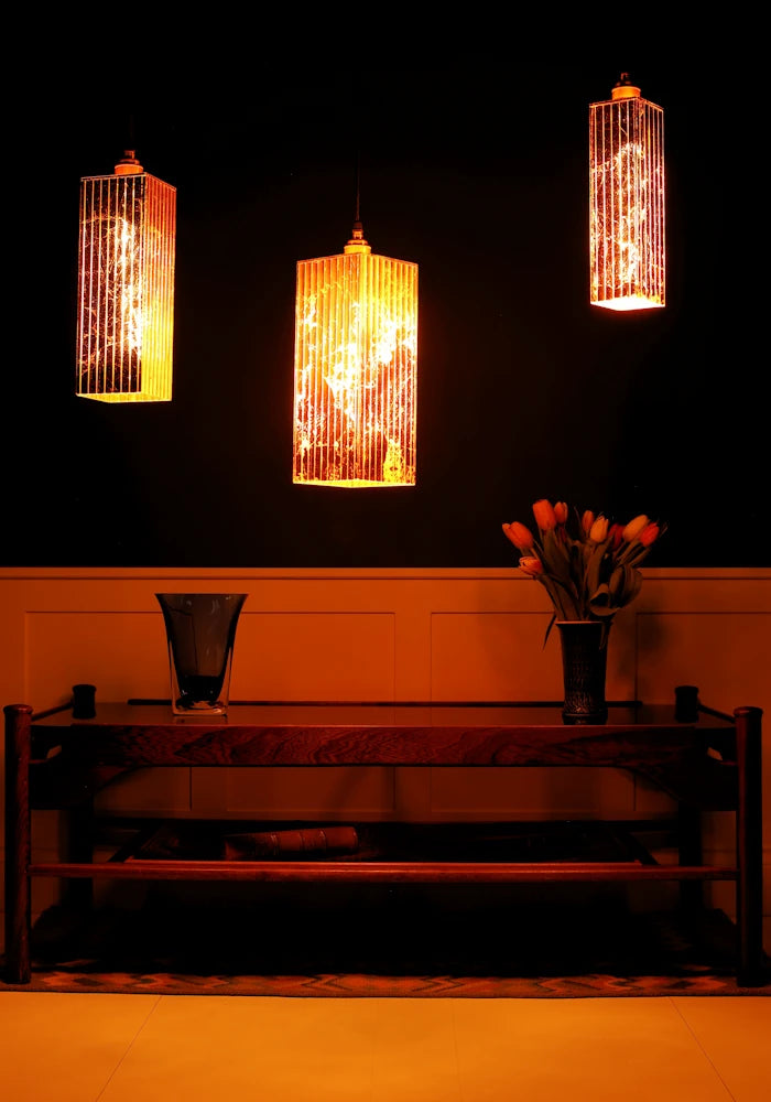 Three illuminated pendant lights hanging above a wooden table with a vase and flowers in a dimly lit room.