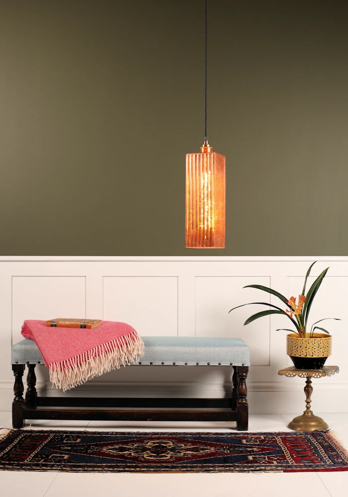 Feature copper leaf pendant light hanging over wooden bench with blanket and plant. White and sage green background.