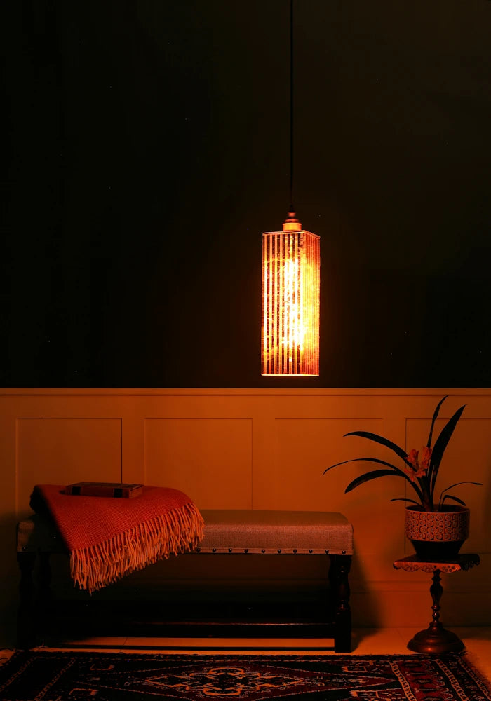 Feature copper leaf pendant light hanging over wooden bench with blanket and plant in darkly lit room