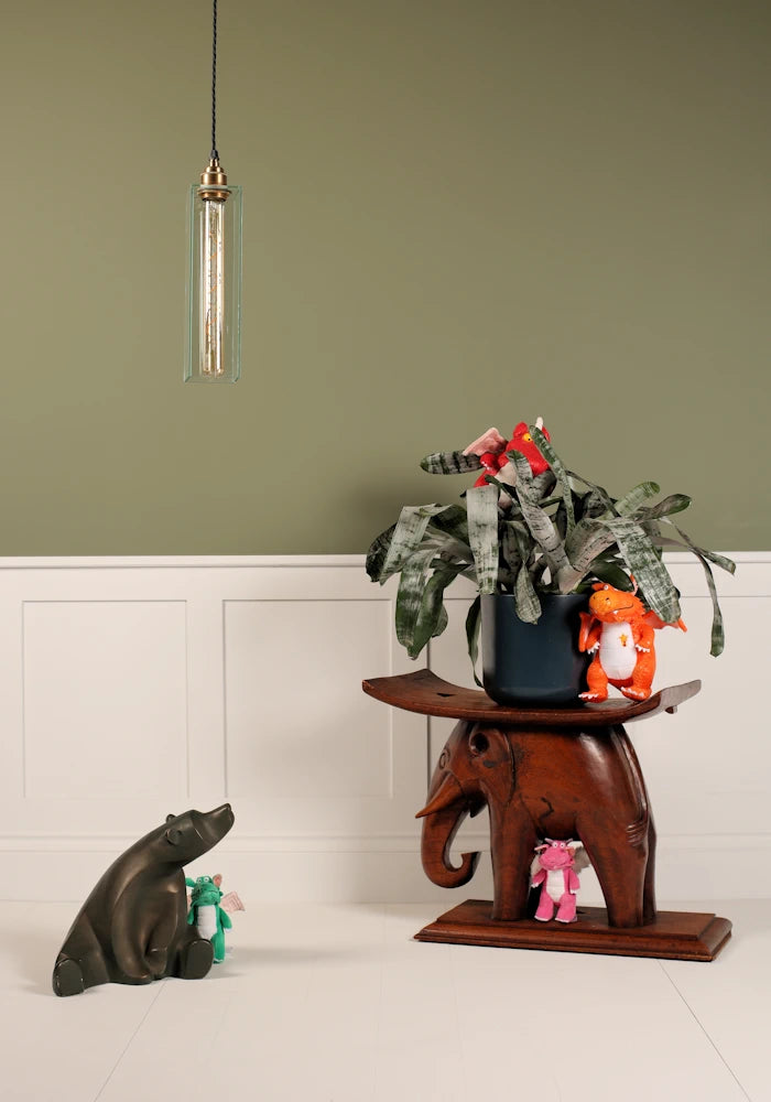 Elegant long clear glass pendant light hanging over small bear statue, wooden carved elephant with pot plant, on sage green and white background 