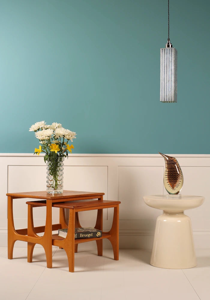 Slim, elegant silver pendant light hanging over ornate vase, nest of coffee tables, flowers on teal background