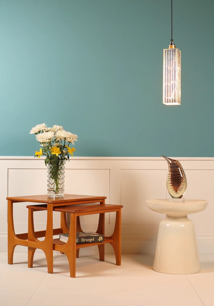 Slim, elegant silver pendant light hanging over ornate vase, nest of coffee tables, flowers on teal background