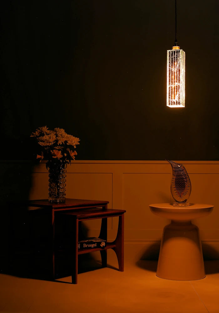 Slim, elegant silver pendant light hanging over ornate vase, nest of coffee tables, flowers in dimly lit room