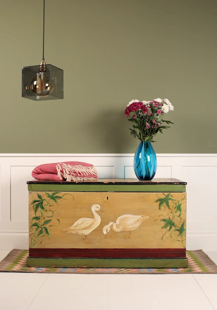 Decorative smoked glass pendant light, ornate chest with swan design on a wooden floor against a green wall.