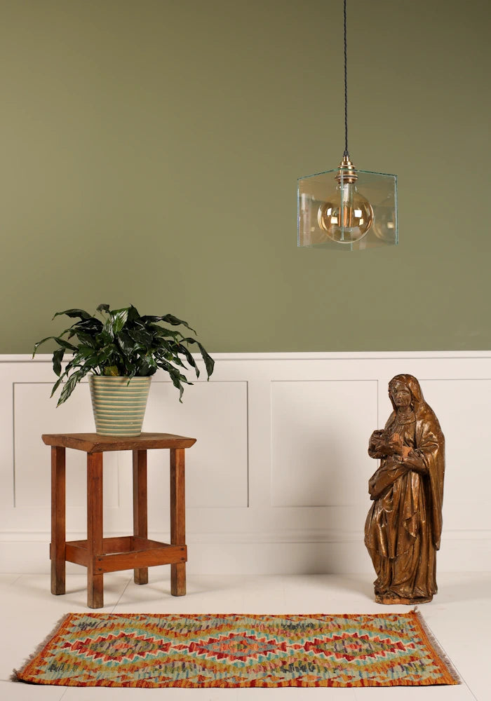 Clear glass cube pendant light, wooden religious statue, pot plant on small table on sage green and white background