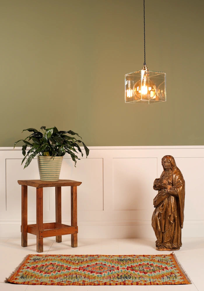 Clear glass cube pendant light, wooden religious statue, pot plant on small table on sage green and white background