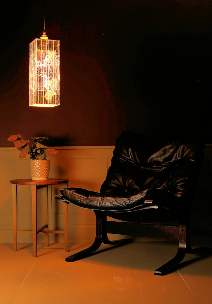 Dark leather chair with a small table and plant under a gold leaf hanging light fixture in a dimly lit room.