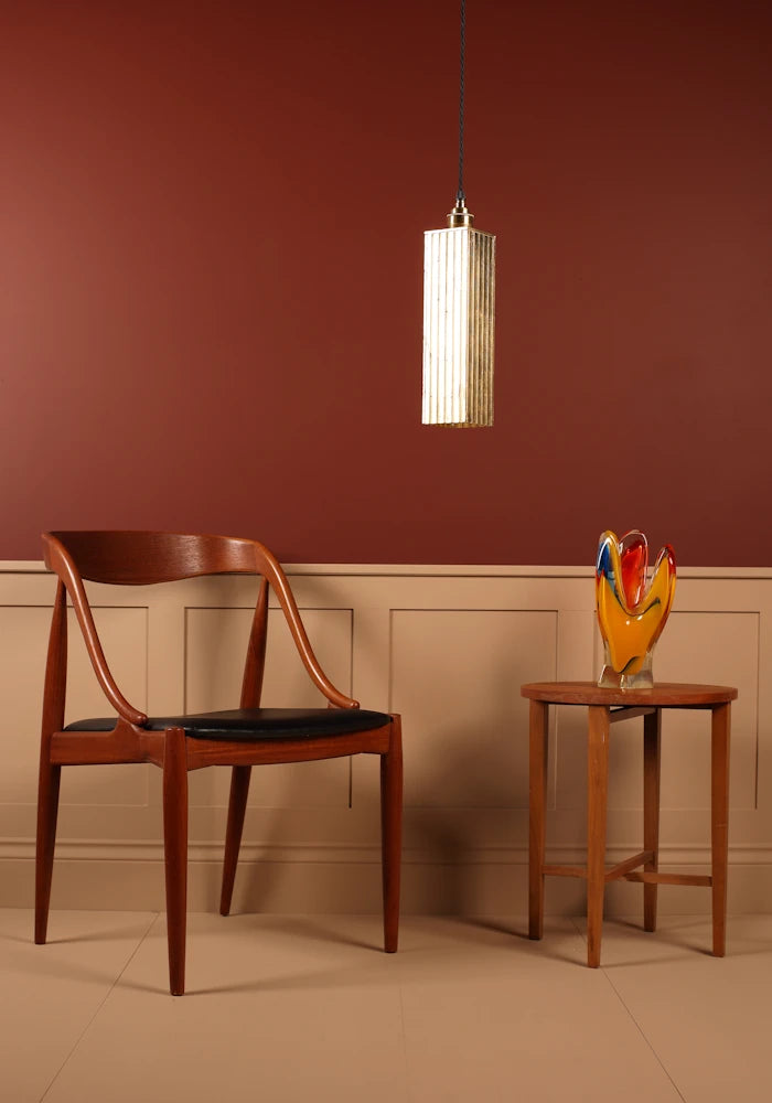 Wooden chair with black seat next to a small table with a gold pendant light and colourful glass sculpture against a red wall.