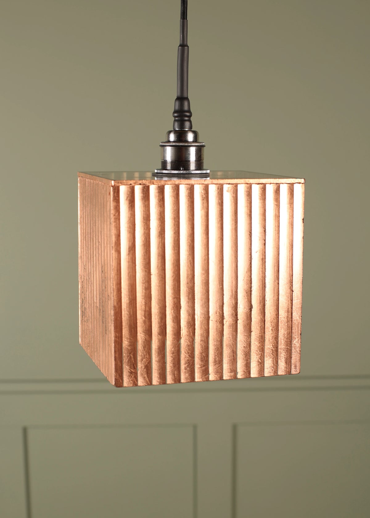 A hanging pendant light with a square, copper-toned fluted shade suspended from a dark metal fixture against a muted green wall.