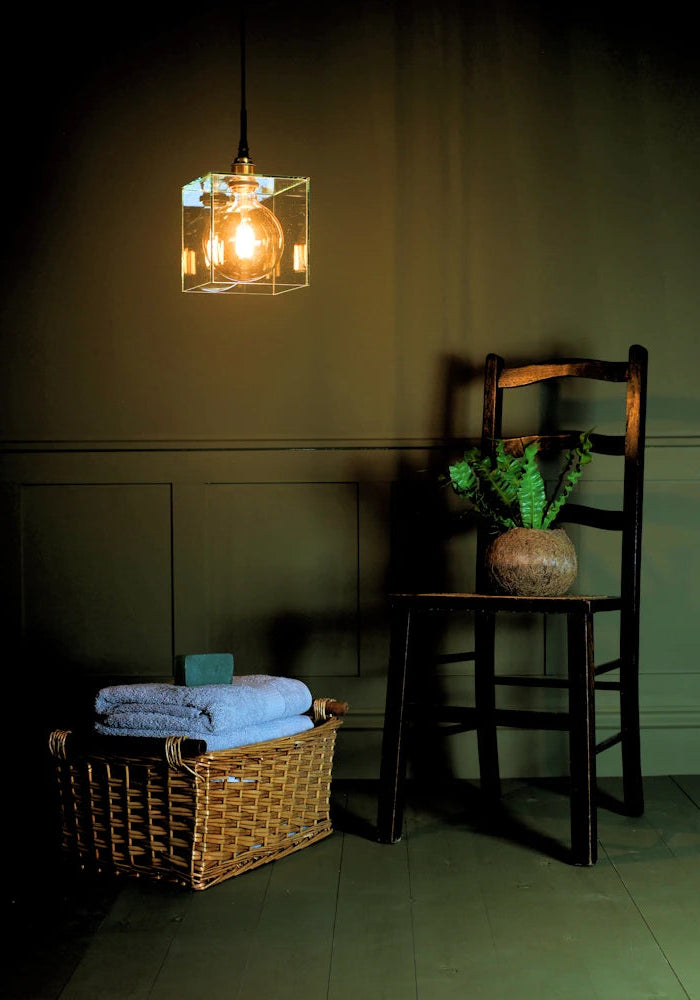 Square clear glass bathroom pendant light casting shadows on wooden chair, plant and towels 