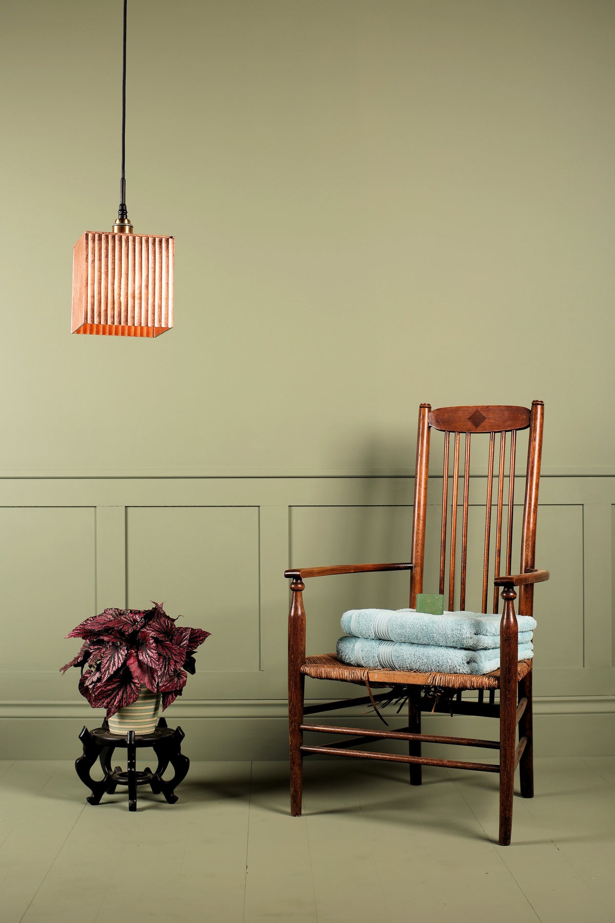 A wooden armchair with a woven seat, styled with folded light blue towels, set against a sage green paneled wall. A hanging copper-toned pendant light and a potted plant on a small stand are visible in the room.