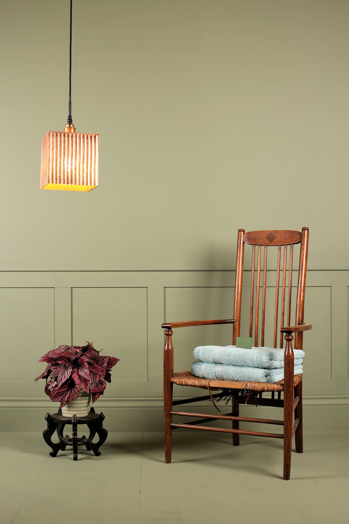 A warm brown wooden armchair with a woven seat, styled with a folded light blue towel, set against a sage green paneled wall. A hanging striped pendant light with a yellow glow and a small potted plant on a stand are positioned to the left.